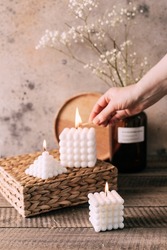 Female hand lights candles. Three white bubble candles on straw stand on wooden background.