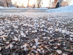 Salt grains on icy sidewalk surface in the winter. Applying salt to keep road clear and people safe in winter weather from ice or snow. Macro view of salt grains with winter scenery in bacground