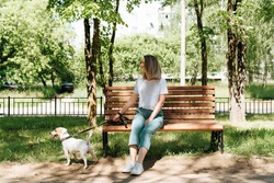 Pretty young woman in stylish clothes resting on a bench while walking the dog. Happy blonde girl and jack russell terrier on a walk in the park on a summer sunny day.