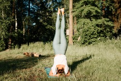 Slender young woman practicing yoga asana headstand back view, outdoors. Sportive girl exercising on a yoga mat in nature in the morning, outside. Healthy active lifestyle concept.