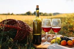 Picnic, romantic evening in nature concept. Bottle of red wine, glasses with drink, grapes, peaches on wooden board, wicker picnic basket on sunset background