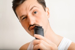Portrait of handsome brutal young man shaving with modern electric razor and looking at camera. Selective focus on trimmer and man's mustache. Men's daily care and beauty concept.