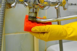 Side view of woman's hand in rubber glove with sponge wiping rusty, old chrome faucet. Close-up housewife cleaning plumbing fixtures from dirt in bathtub indoors. Household cleaning, bathroom washing.