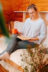Literary hobby, cozy intellectual leisure concept. Adult caucasian woman wearing glasses reading book with mug of coffee while sitting on sofa at home.