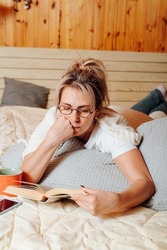 Adult caucasian woman in glasses reading book in concentration lying on bed in bedroom. Middle-aged woman and literary hobby, intellectual leisure. Vertical photo.