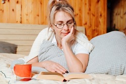 Reading book, literary hobby, intellectual leisure concept. Adult caucasian woman in glasses resting lying on bed with book and mug of coffee or tea in bedroom, indoors.