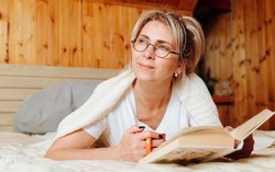 Adult caucasian woman wrapped in plaid wearing glasses and holding mug of coffee reading book, relaxing on bed and looking away, indoors. Literary hobby, intellectual leisure.
