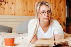 Portrait of middle aged caucasian woman wearing glasses and wrapped in knitted blanket resting on bed with mug of coffee or tea and reading book, indoors. Literary hobby, cozy leisure.