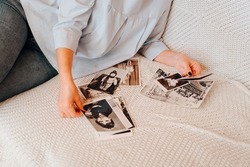 Top view of woman looking at her retro photographs, nostalgic for childhood and youth, sitting on couch at home. Close-up of female hands holding black and white film photos. Selective focus.