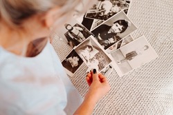 Close-up caucasian woman looking retro photographs of childhood and youth, nostalgic sitting on sofa at home. Back view of female hands holding black and white film photos. Selective focus on pictures