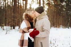 Young hipster couple in love hugs and kisses in winter forest, outdoors. Caucasian family covered with blanket with cups of hot drink together on walk in vacation. Romantic date, valentine's day.
