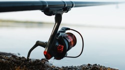 Spinning rod on shore near water outdoors, close-up of steel red reel. Fishing hobby, sport and leisure concept. Selective soft focus.