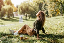 Young caucasian woman resting carefree, relaxing on lawn on sunny day. Side view hipster woman with closed eyes enjoying calmness and freedom during coffee break while sitting on grass outdoors.