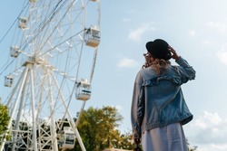 Young curly hipster woman holding hat and looking at ferris wheel in city amusement park on sunny day, outdoors. Back view of caucasian woman enjoying fair view, lifestyle and leisure.