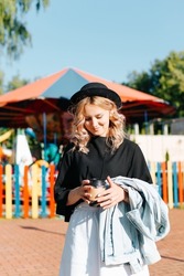 Young curly woman with cup of coffee standing in front of carousel in an amusement park and looking down. Pretty woman in round hat enjoying festival, outdoor fair.