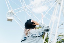 Young hipster woman in hat and denim jacket looking at ferris wheel in city amusement park on sunny day. Woman enjoying view at fair, outdoors. Close-up, back view.