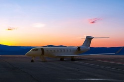 Modern white private jet with an opened gangway at the airport apron on the background of a picturesque sunset