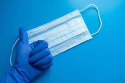 Hand in medical glove thumb up protective mask on blue background. A typical 3-layer surgical mask for covering the mouth and nose. The concept of protection against bacteria and viruses.