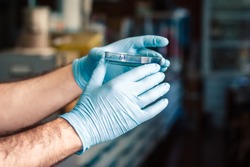 Hands in blue rubber protective gloves holding a Petri dish with a chemical sample in laboratory, with blurred background