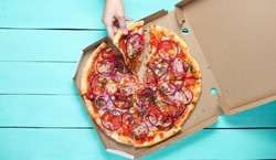 The hand takes a piece of pizza in box on blue concrete table. Top view