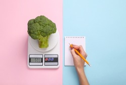 Diet, weight loss, calorie counting concept. Broccoli on a kitchen scale, a hand writes information in a notebook on a blue-pink pastel background. Top view