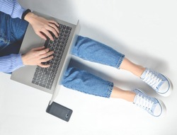 The concept of blogging, working on a laptop. Using a laptop sitting on a white floor. Modern technologies. Female legs in jeans and sneakers. Top view. Flat lay.