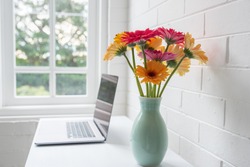 Closeup of pink and orange gerbera daisies in green vase on white home office desk with laptop and window in background (selective focus)
