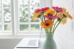 Closeup of yellow and pink gerbera daisies in green vase on desk with laptop next to window (selective focus)