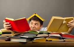 Middle-aged shocked man littered with books, reading, holding two books in hands on a pile of books on table. Concept too much reading. Get lost in the information. A lot of knowledge