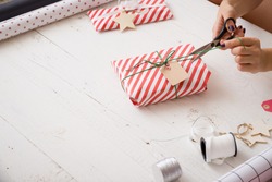 Young woman's hands wrapping Christmas gift in striped paper and decorating it with and twine and tag on white wooden background. Girl wrapping presents for the party.
