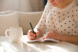Woman sitting at the table, writing in the notebook and drinking coffee in nice light home interior. Working at home. Freelancer. writing down ideas. indoors.