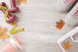 Fall season physique transformation. Top view photo of dumbbells, floor scales, tape measure, trendy shoes, bottle, dry maple leaves on light wooden background with advertising zone