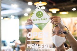 Business owner Asian woman wear protective face mask ppe hanging open sign at her restaurant / café, open again after lock down due to outbreak of coronavirus covid-19