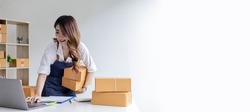 Asian woman holding parcel boxes and checking orders from laptop, she owns an online store, she packs and ships through a private transport company. Online selling and online shopping concepts.