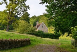 Scenic view of a stone cottage house in a green field with green leafy trees in rural England