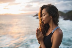 Young woman praying and meditating alone at sunset with beautiful ocean and mountain view. Self-analysis and soul-searching. Spiritual and emotional concept. Introspection and soul healing.