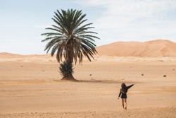 Woman walking in beautiful desert with sand dunes and one lonely palm. View from behind. Freedom and travel concept.