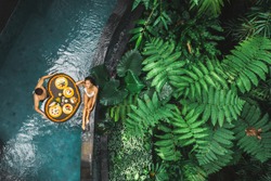 Travel happy couple in love eating floating breakfast in jungle swimming pool. Awakening in morning. Black rattan tray in heart shape, Valentines day or honeymoon surprise, view from above.