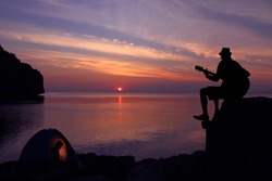 Silhouette couple camping and playing a guitar on the beach with sunset sky background.