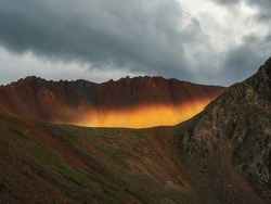 Caldera is flooded with sunlight. Landscape with sunlit wide sharp mountain ridge between mountain tops. Colorful mountain scenery with large sharp rocks on ridge top in sunlight under cloudy sky.