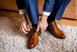 A man ties up his shoelaces on his brown shoes in the room. Blue suit and patent leather shoes. 