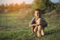 Happy Asian Child (poor kid) smiling and hugging teddy bear, sitting on grass. Teddy Bear and child are best friends. Friendship and homeless people concept.