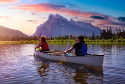 Couple adventurous friends are canoeing in a lake surrounded by the Canadian Mountains. Colorful Sunrise Sky Art Render. Taken in Vermilion Lakes, Banff, Alberta, Canada.