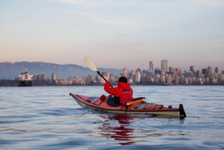 Adventure Man on a Sea Kayak is paddline during a vibrant winter sunset with Downtown City Skyline in the background. Taken in Vancouver, British Columbia, Canada.