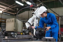 Female automation engineer wear a blue uniform with helmet safety inspection control a robot arm welding machine with a remote system in an industrial factory. Artificial intelligence concept.