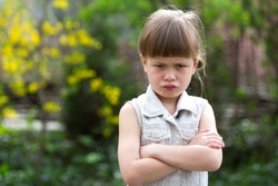 Pretty funny moody little blond preschool girl in white sleeveless dress looks into camera feeling angry and unsatisfied on blurred summer background. Children tantrum concept.