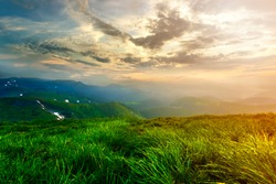 Wide summer mountain view at sunrise. Glowing orange sun raising in blue cloudy sky over green grassy hill soft grass and distant mountain range covered with morning mist. Beauty of nature concept.