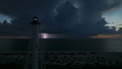 White tall lighthouse on sea shore with blinking light at stormy night for commercial vessels navigation. Thunderstorm with lightnings over ocean water posing danger for ships