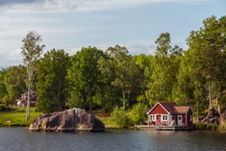 View on red holiday cabin by a lake in Stockholm archipelago, Sweden. Wooden cottage, sauna on shore. Tiny house near the water. Rocky small island, islet in water. Buildings surrounded by green trees