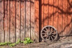 Old wooden wagon wheel reclining on red wooden wall. Wheel with boardwalk wall on background and cobbles on foreground. Place for your text. Wheel leaning up against wall. Sunny day. Skansen, Sweden